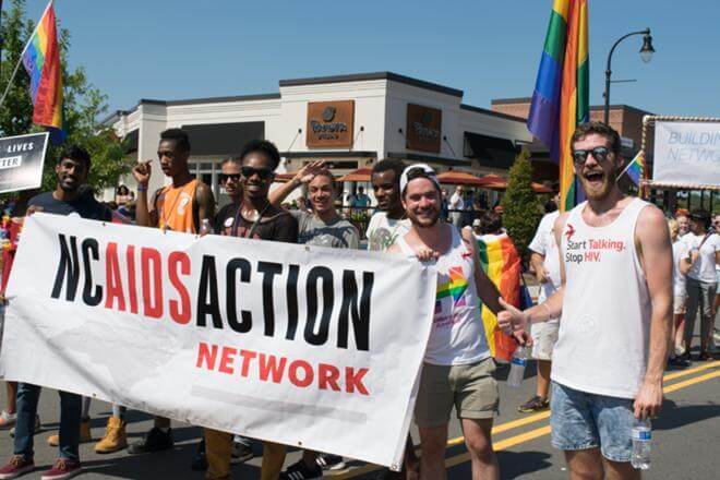 A group of eight people in a Pride parade smiling and waving at the camera. They are holding a large banner that says "NC AIDS Action Network."