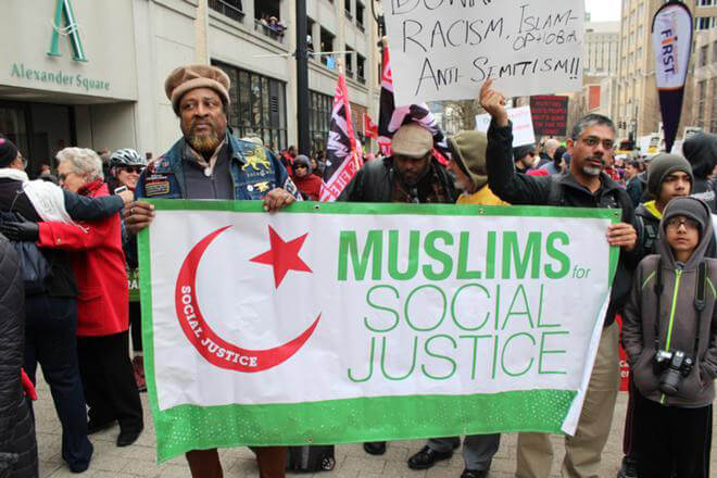 A crowd of people outside, two of whom are holding a large green, white, and red banner that says "Muslims for Social Justice." One of them also holds a paper sign. The top of the sign is out of frame, but the bottom says "racism, Islamophobia, anti-Semitism!!!"