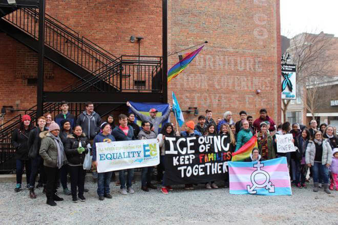 A group of around 35 people standing in front of a brick building. People in the front hold a banner with the Equality NC logo, a banner that says "ICE out of NC, keep families together", a transgender flag with a transgender symbol on it, and a sign that says "youth power!"